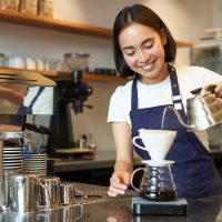 Cute brunette girl barista, cafe staff pouring water from kettle and brewing filter coffee behind counter, preparing order.